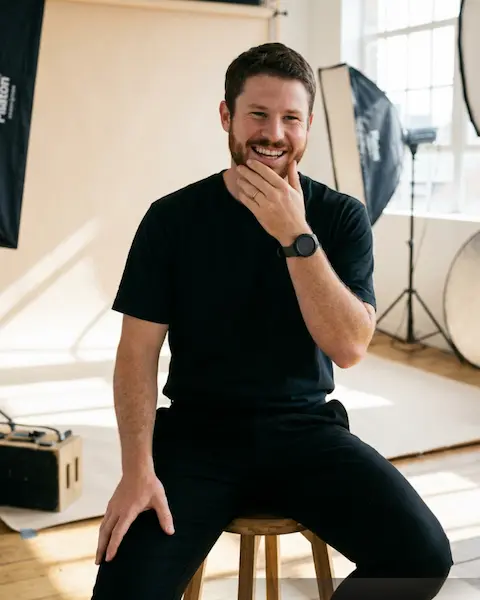 A smiling portrait of a man with a beard and a gold wedding ring, sitting on a wooden stool. He is in a photography studio with softbox lights, backdrops, and other studio equipment visible, and daylight comes through a window.
