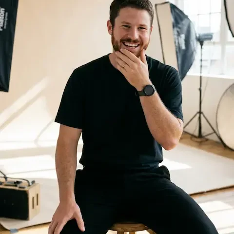 A smiling portrait of a man with a beard and a gold wedding ring, sitting on a wooden stool. He is in a photography studio with softbox lights, backdrops, and other studio equipment visible, and daylight comes through a window.